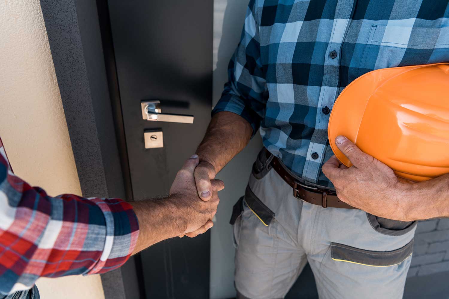 Builder in blue checkered shirt holding an orange hat shakes hands with someone at their home